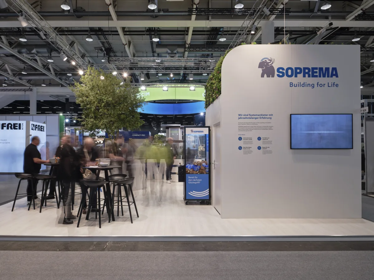 Seitenansicht des Soprema Messestands auf der Swissbau 2026 mit Lounge-Bereich, Stehtischen, Bildschirm und Soprema-Logo an der Rückwand