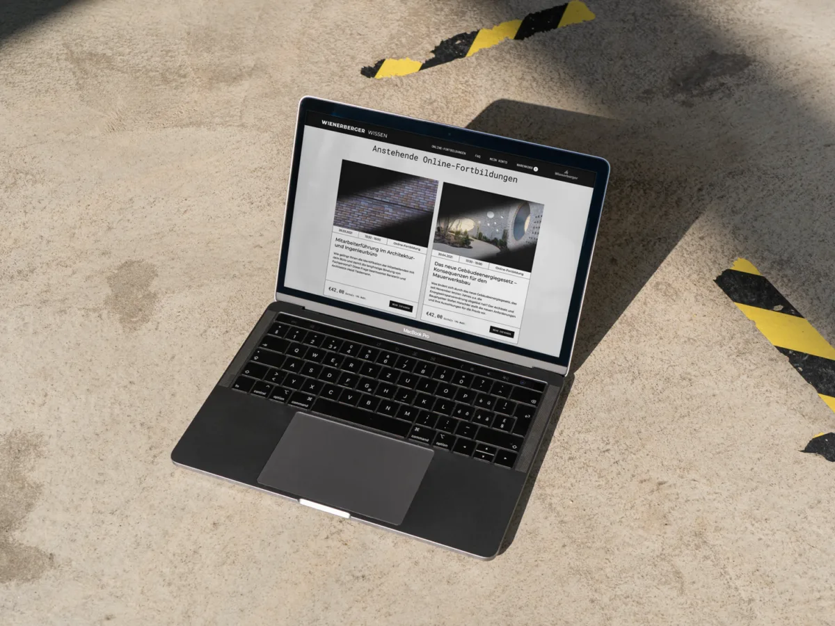MacBook auf Betonboden zeigt die Kursübersicht von wienerberger-wissen.de mit gelb-schwarzem Absperrband im Hintergrund