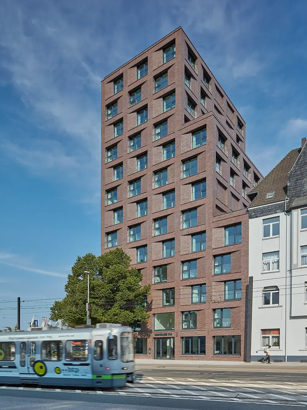 Modernes Hochhaus mit skulpturalem Eingang in der Stadt Hannover, im Vordergrund fährt eine Straßenbahn vorbei.