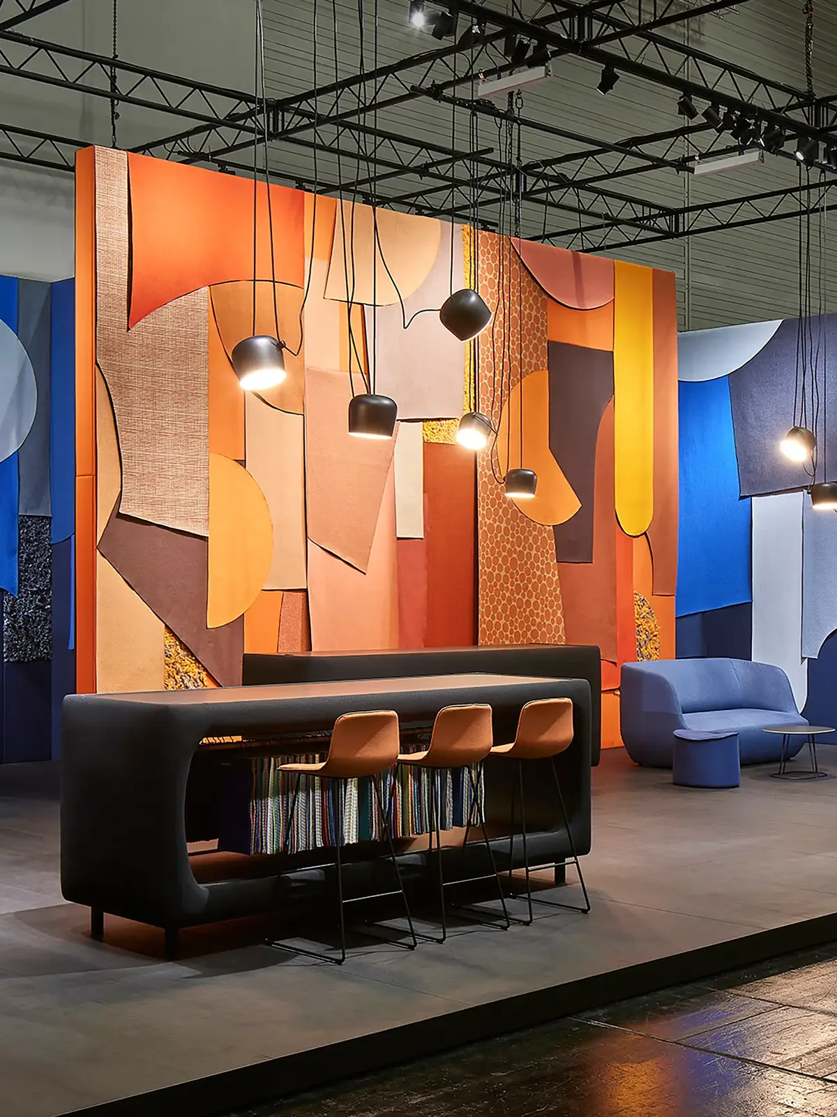 Messestand von Camira auf der ORGATEC 2022: farbenfrohe Wandgestaltung mit geometrischen Textilflächen in Orange-, Braun- und Rottönen, kombiniert mit modernen Möbeln, Barhockern und Pendelleuchten.